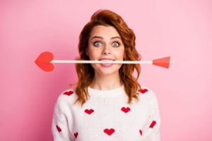 A woman wearing a white sweater with red hearts is holding an arrow with a heart at the tip between her teeth