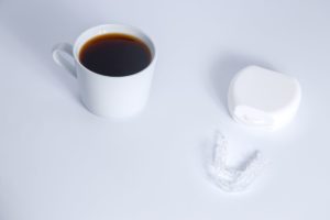Cup of coffee sitting next to clear aligner trays and their carrying case on a white background